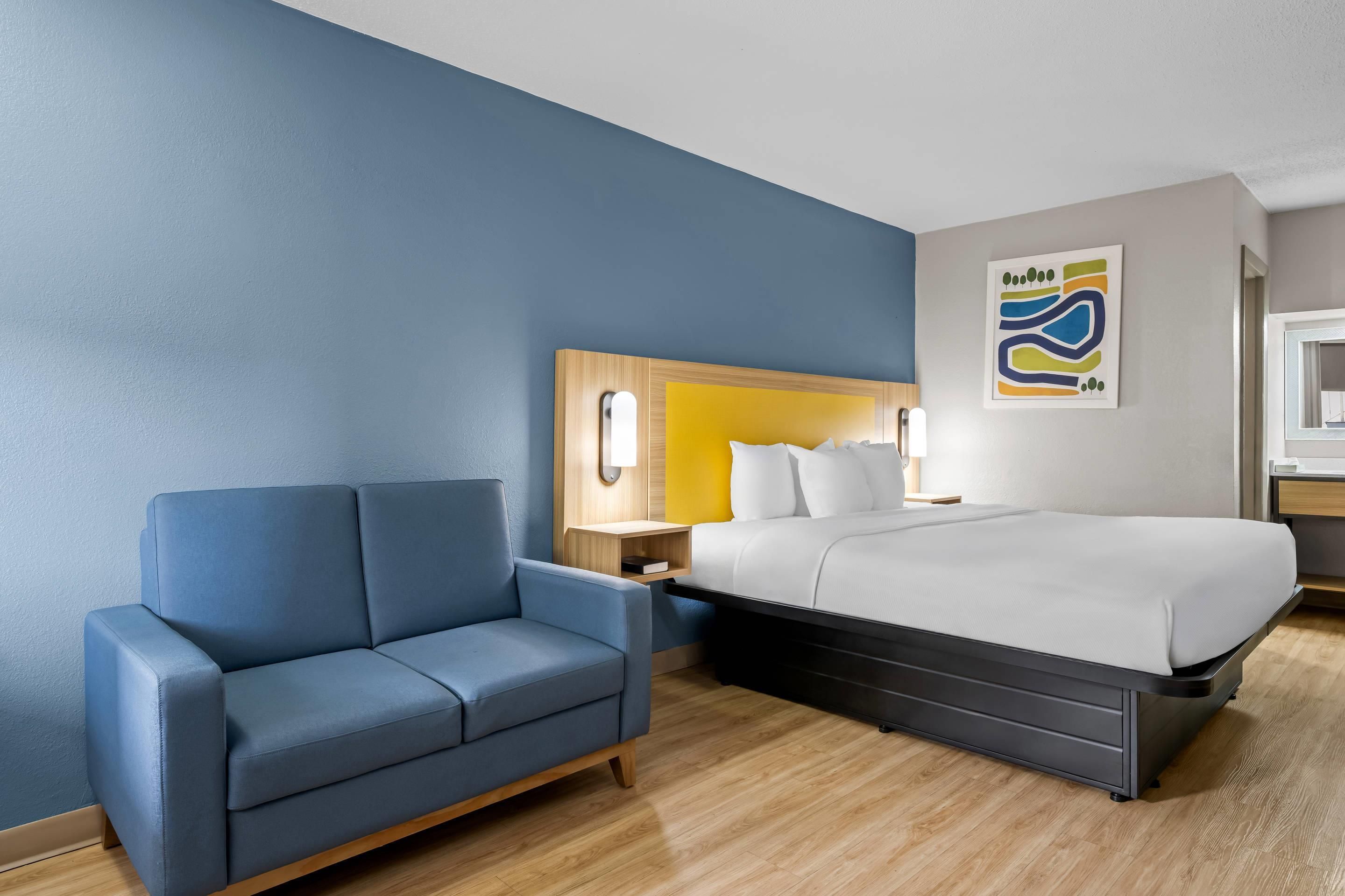 Una habitación de hotel con cama, un sofá azul, una mesita de noche de madera y una obra de arte colgada en la pared.