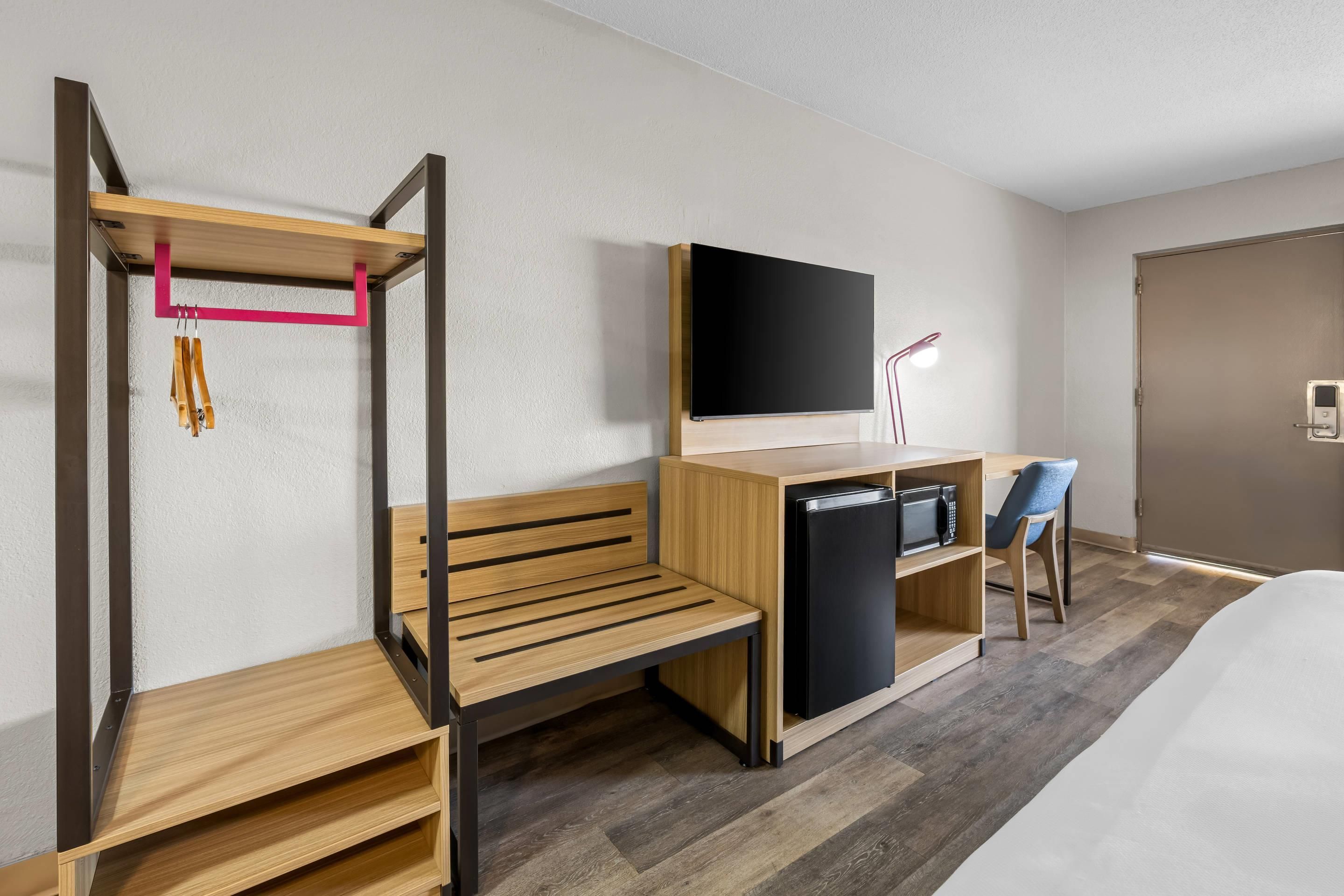 A compact hotel room with a bed, a desk with a flat-screen TV, a small fridge, and a bench.