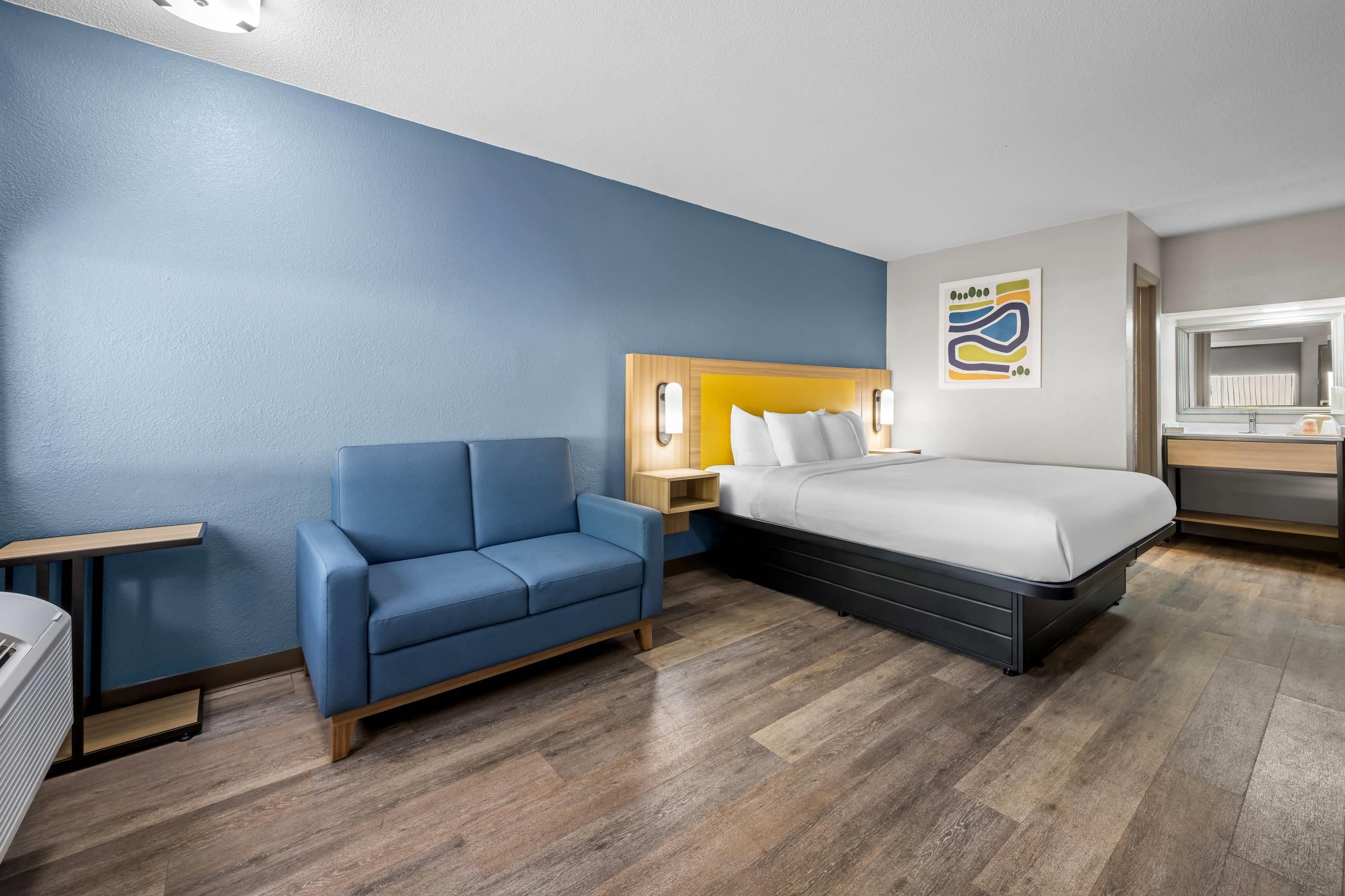 Habitación de hotel con cama, un sillón azul, una mesita de noche y una obra de arte colgada en la pared.