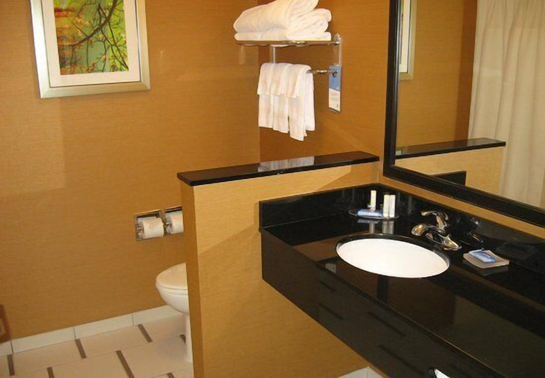A bathroom with a black countertop, white sink, and a mirror. There is a towel rack with folded towels and a framed picture on the wall.