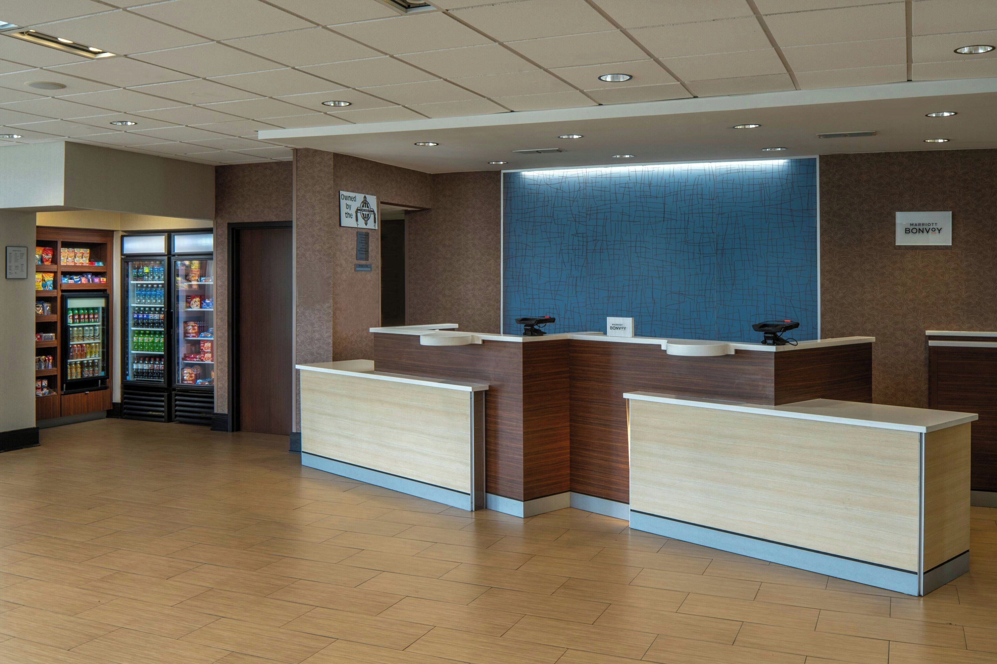 A hotel reception area with a modern design, featuring a reception desk, a blue textured wall, and a vending machine with snacks and drinks.