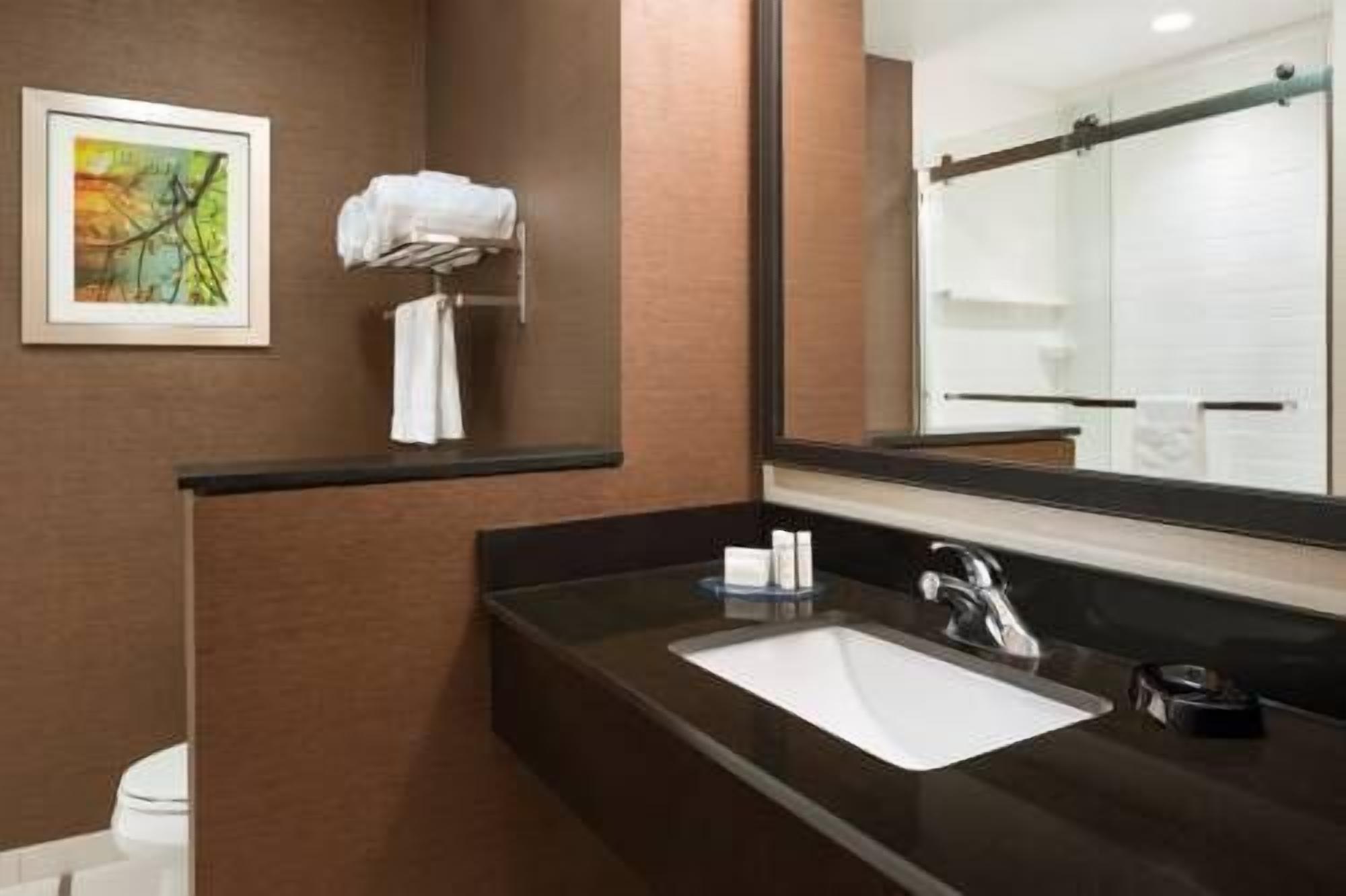 A modern bathroom with a dark brown countertop, a white sink, and a large mirror. There is a towel rack with a white towel and a framed picture on the wall.