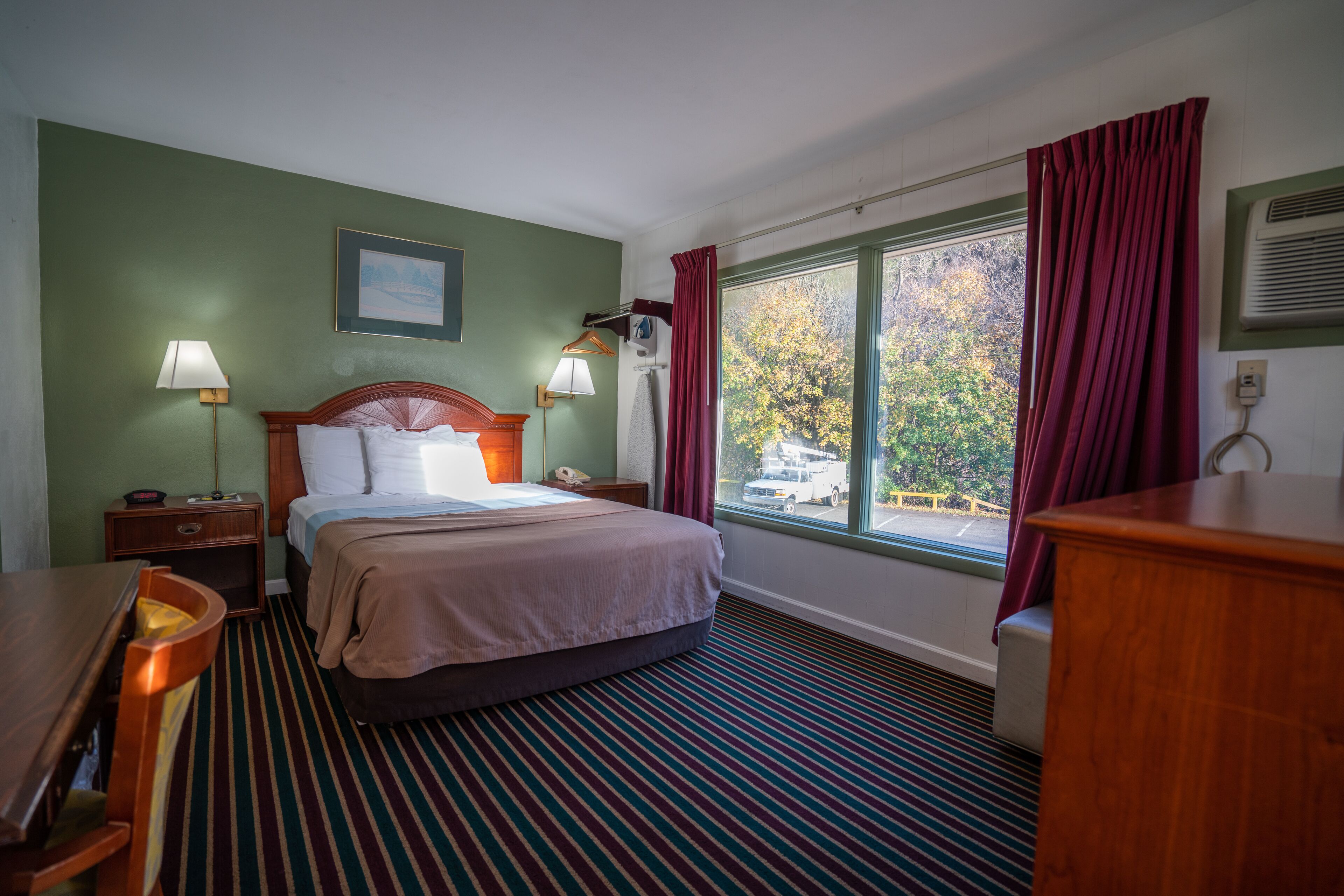 Ein Hotelzimmer mit Bett, Nachttischen, Lampen, einem Schreibtisch und einem fensterbepflanzten Fenster.