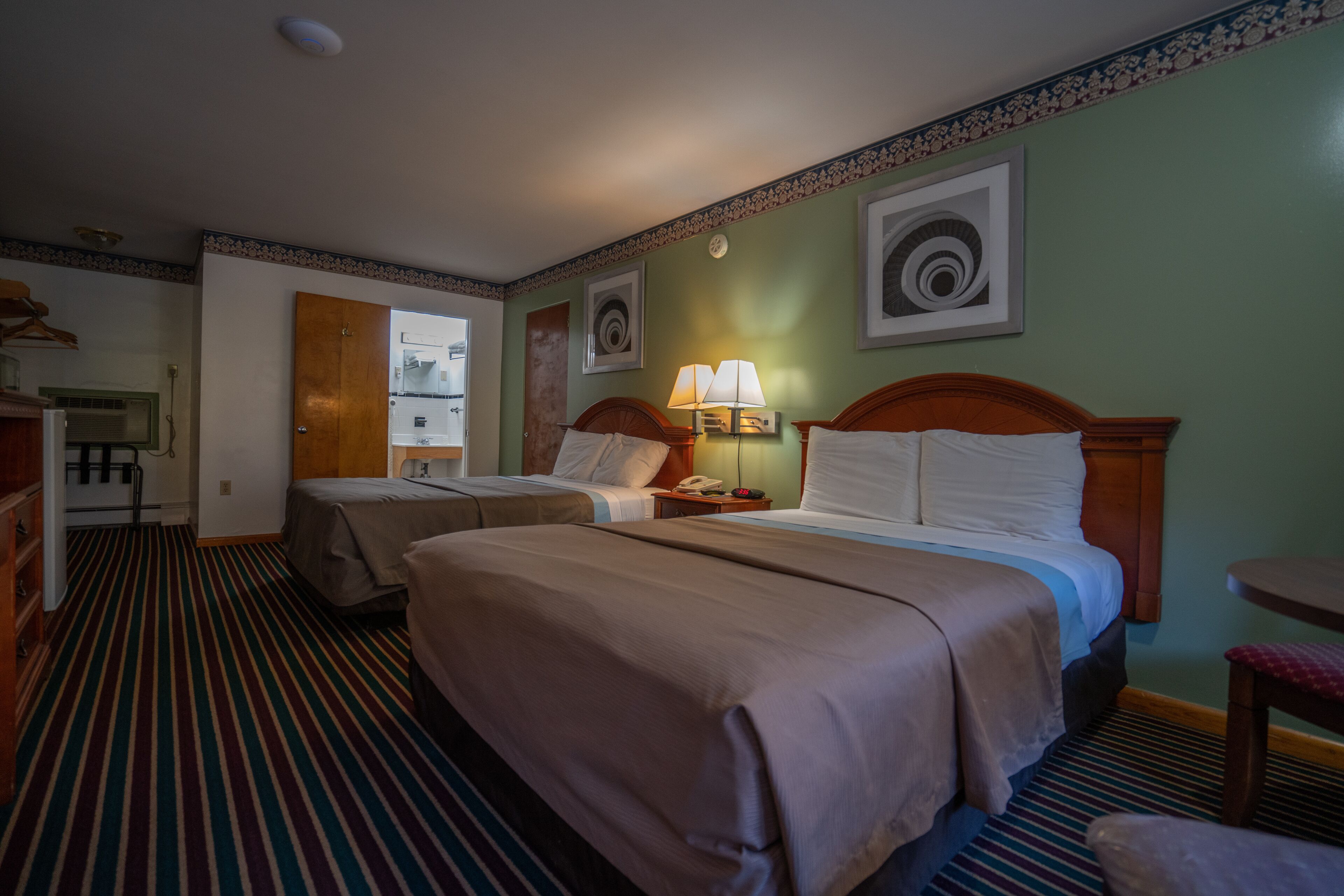 A hotel room with two beds, a small table, a chair, a desk with a lamp, and a door leading to a bathroom.