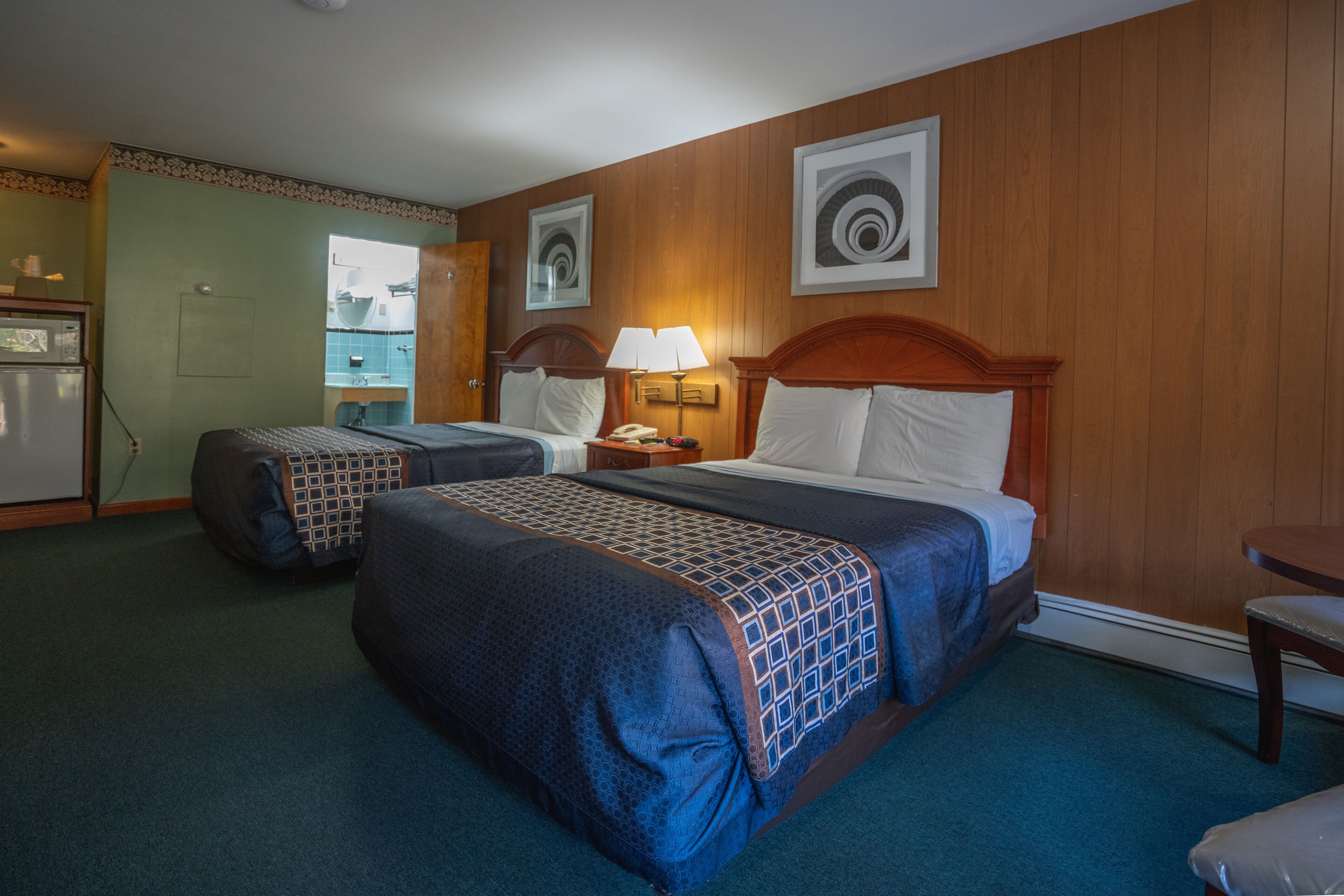 A hotel room with two beds, a desk, a chair, a lamp, and a refrigerator.