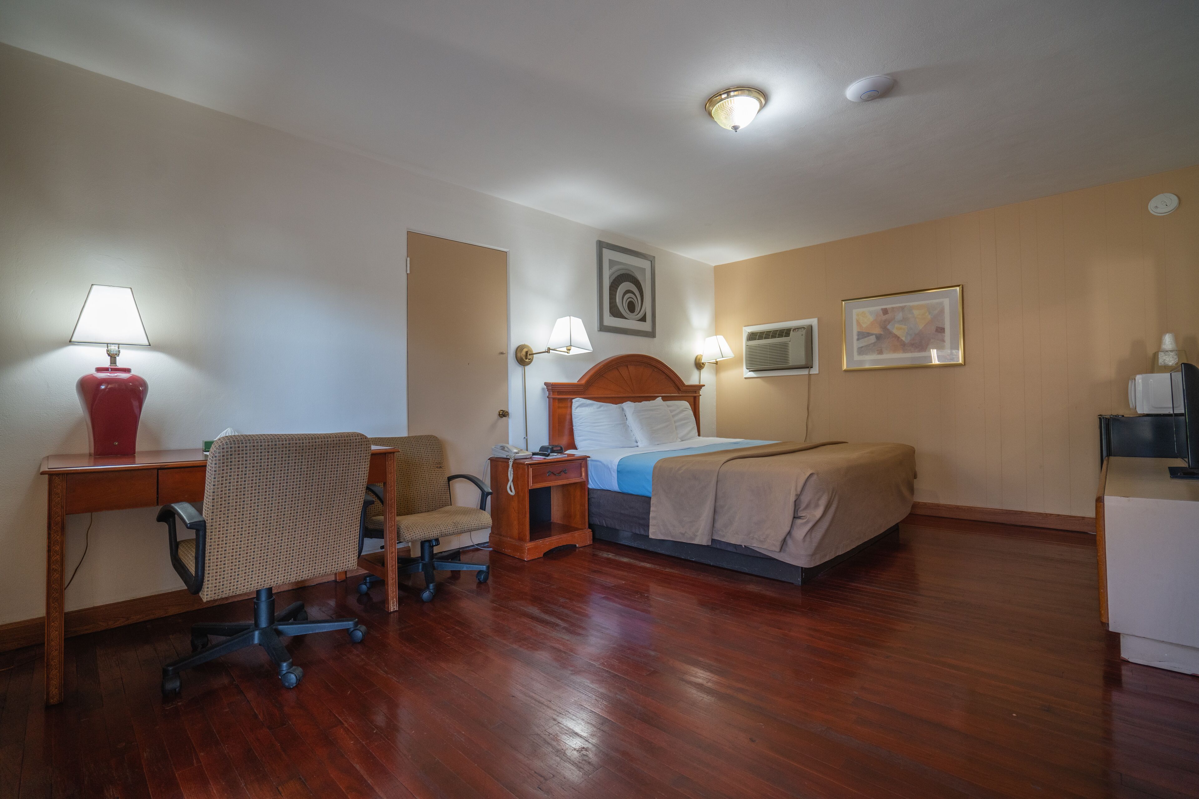 Hotelkamer met een houten bureau, stoel, nachtkastje, lamp, bed en een wandairconditioner.