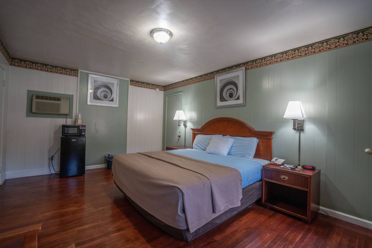 Un dormitorio ordenado con una cama de madera, lámparas de noche, una mesita de noche, un microondas y cuadros en las paredes.