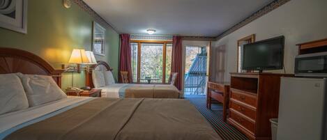 A hotel room with two beds, a television, a dresser, and a balcony with a view of trees.