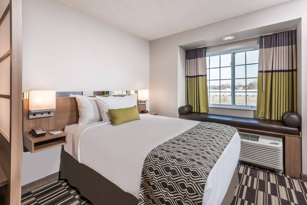 A hotel room with a large bed, bedside tables, a window with curtains, and a built-in heater.