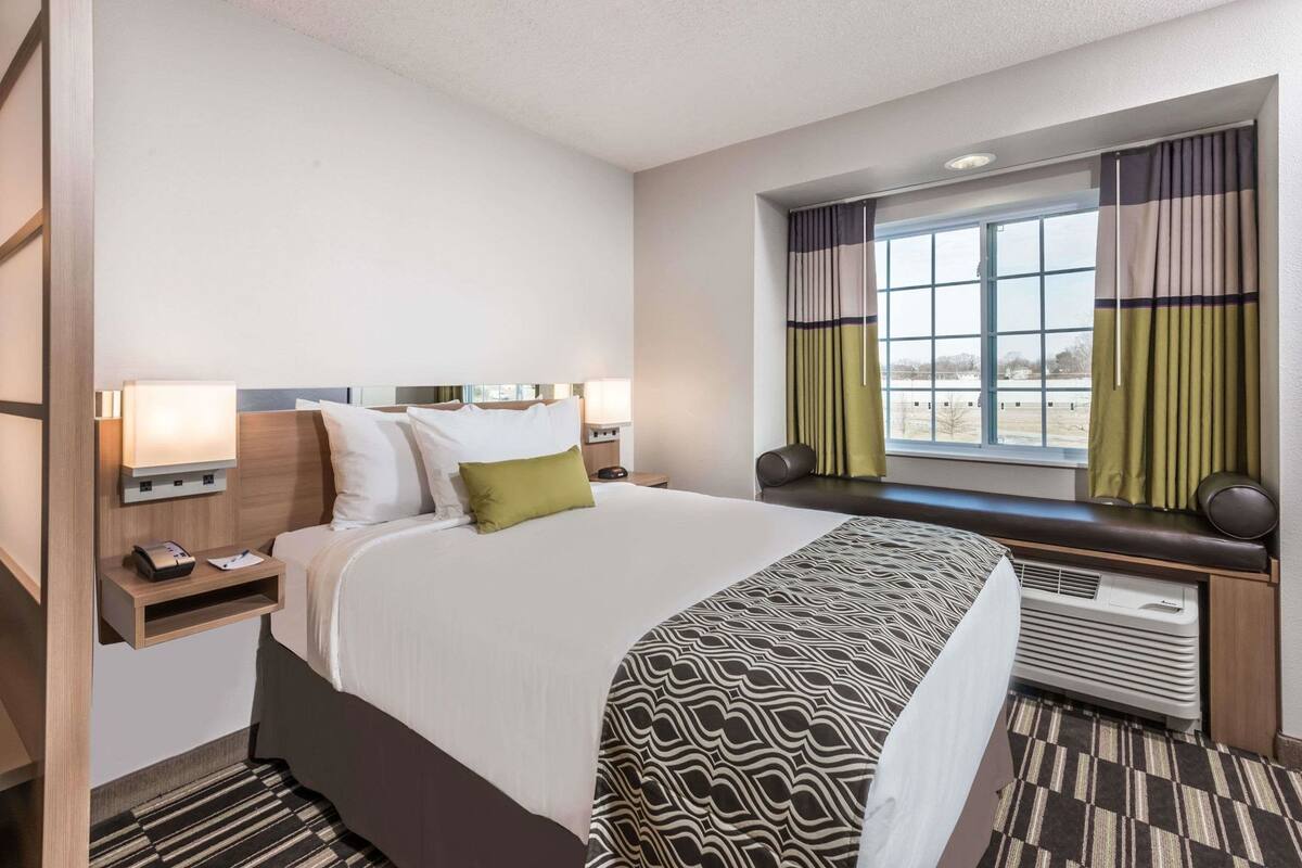 A hotel room with a bed, bedside tables, a window with curtains, a radiator, and a wall-mounted TV.