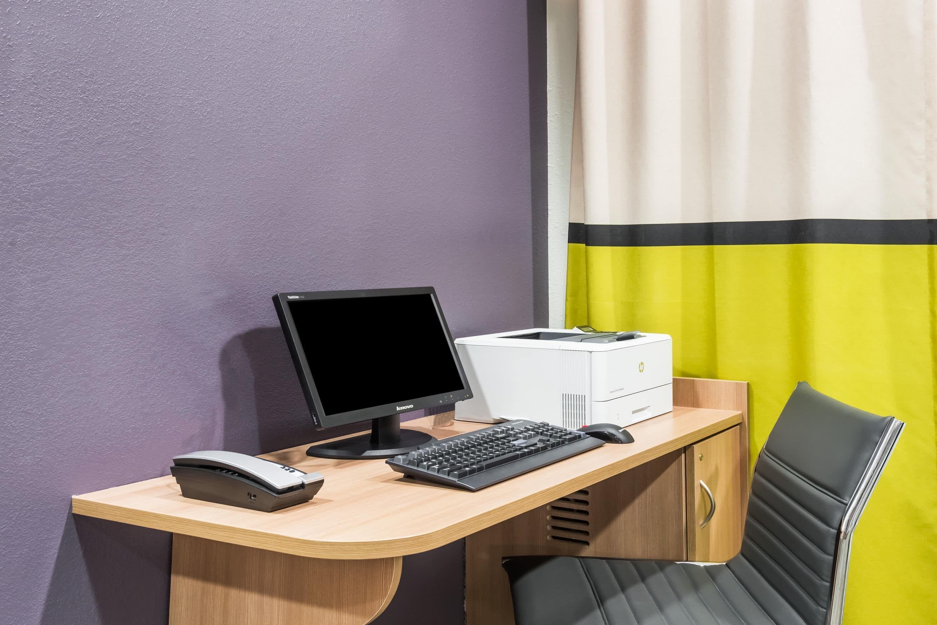 Un espacio de trabajo con una computadora, teclado e impresora sobre un escritorio, una silla y una pared morada.