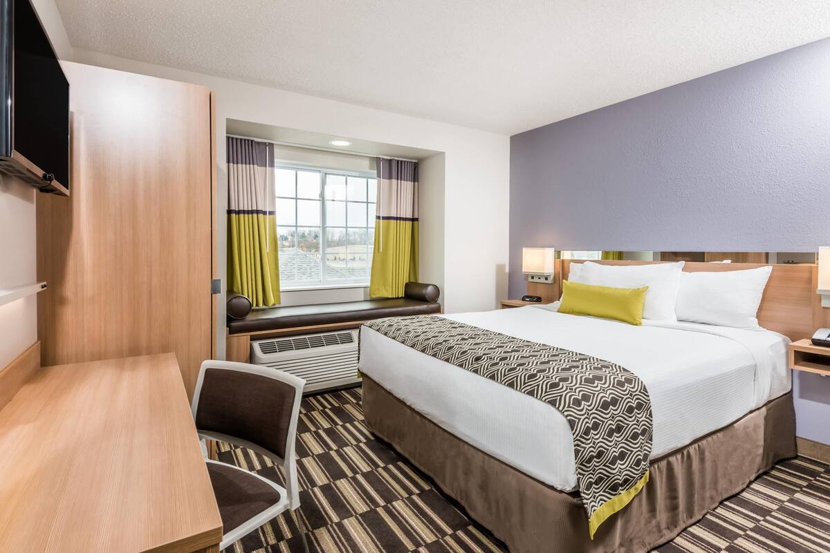 A hotel room with a bed, a desk, a chair, a TV, a window with curtains, and a nightstand with a lamp.