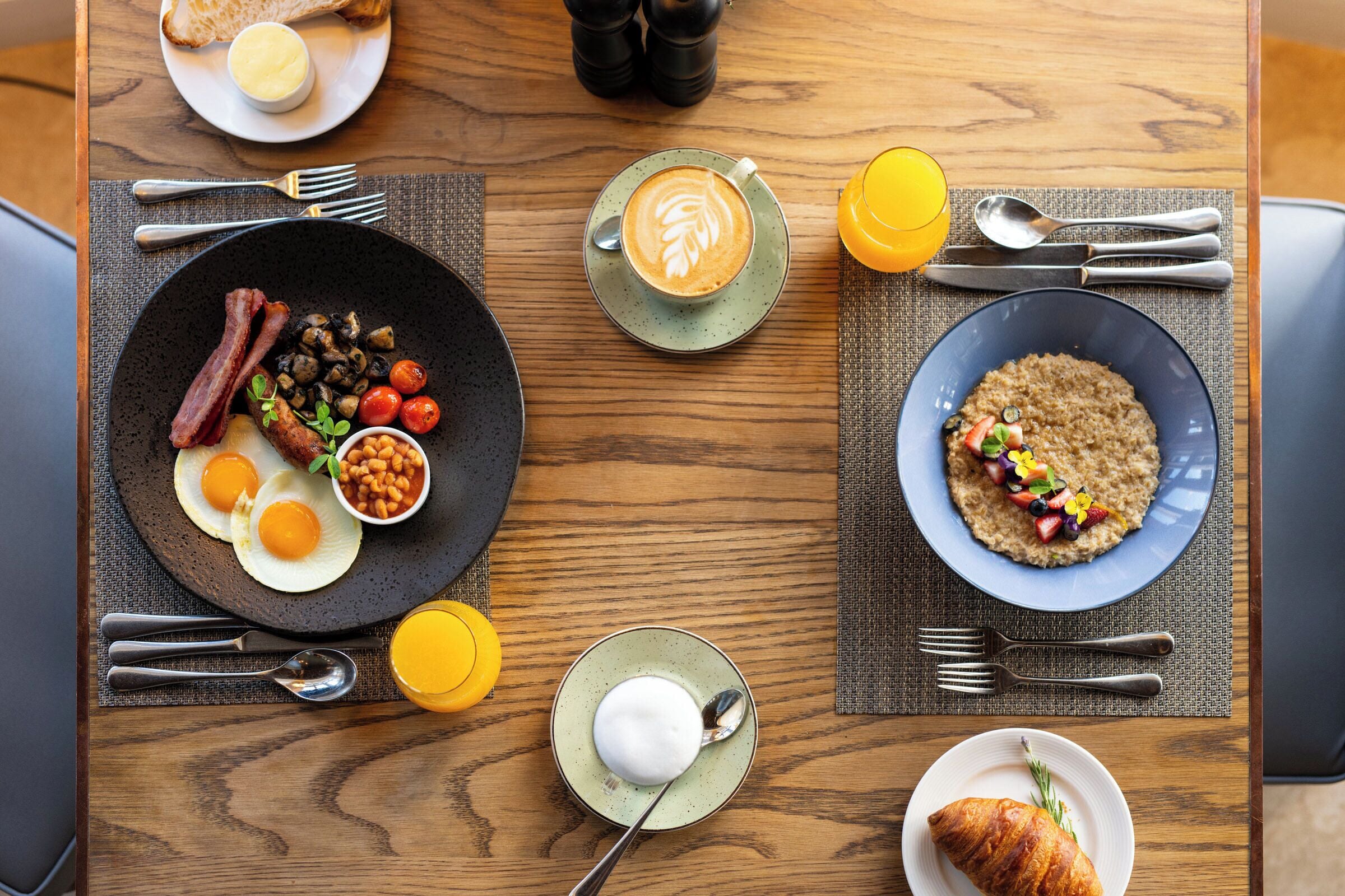 A breakfast setting with a plate of eggs, bacon, mushrooms, and beans, a cup of coffee, a bowl of oatmeal with fruits, and a croissant on a wooden table.