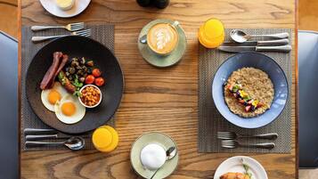 A breakfast setting with a plate of eggs, bacon, mushrooms, and beans, a cup of coffee, a bowl of oatmeal with fruits, and a croissant on a wooden table.