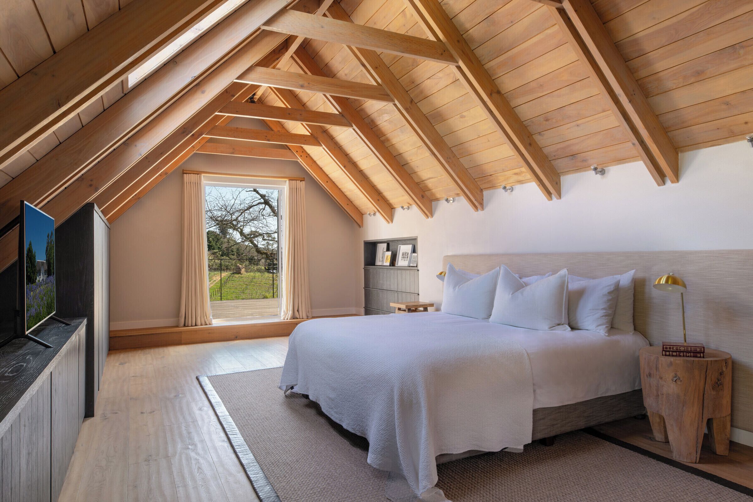 A bedroom with a sloped ceiling, a bed, a bedside table with a lamp, a TV, and a window with curtains.