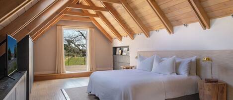 A bedroom with a sloped ceiling, a bed, a bedside table with a lamp, a TV, and a window with curtains.