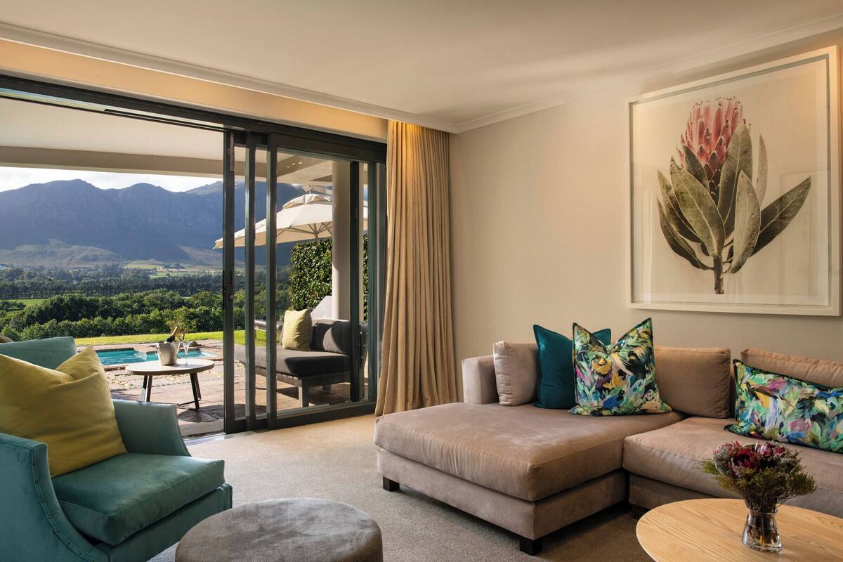 A modern living room with a large sectional sofa, a small round table, and a view of a pool and mountains through large sliding glass doors.
