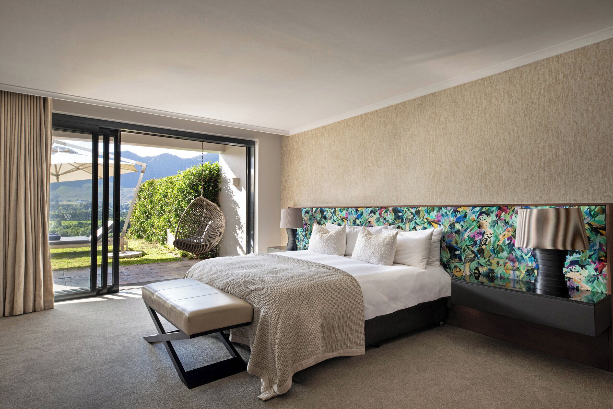 Ein modernes Hotelzimmer mit einem großen Bett, einer Bank, Blick ins Freie und einem farbenfrohen Kopfteil.