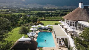 A swimming pool with a thatched-roof building, surrounded by lush greenery and mountains in the background.