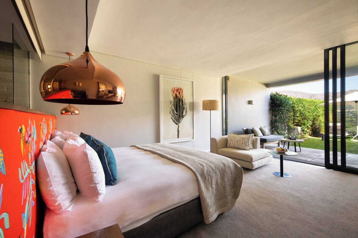 A modern bedroom with a large bed, a hanging pendant light, a sofa, and a view of the outdoors.