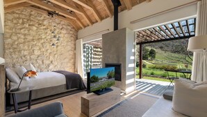 A bedroom with a stone wall, a bed, a TV, and a view of a garden. - Mont Rochelle Hotel & Vineyard (Franschhoek)