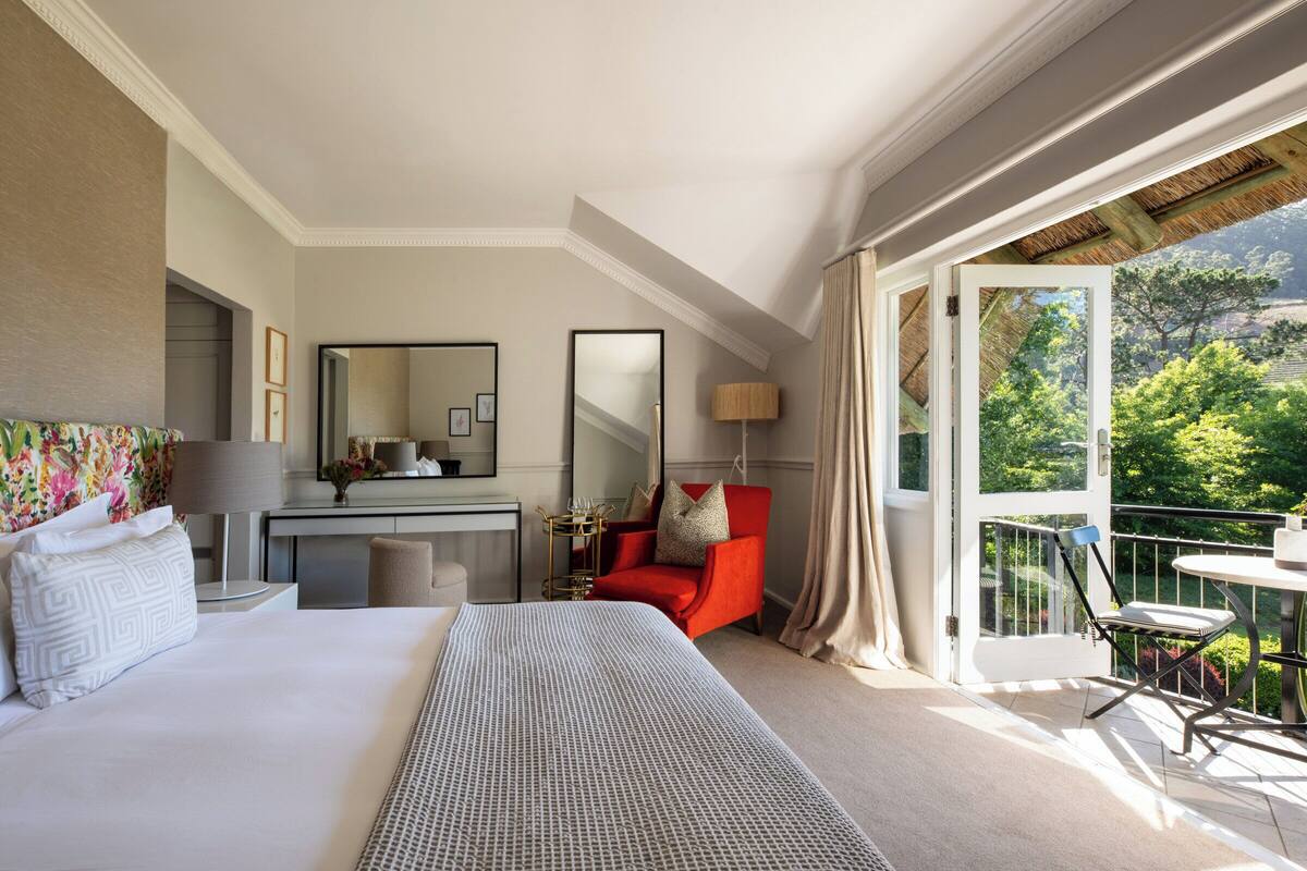 A hotel room with a large bed, a desk, a red armchair, and a balcony with a view of greenery.