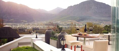 A terrace with a table and chairs, a bottle of MIKO wine, and a view of a valley with mountains.