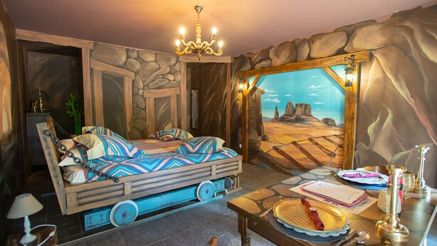 A room with a bed, a table with a plate and utensils, a chandelier, and a mural of a desert scene. - Western City (Chaudfontaine)