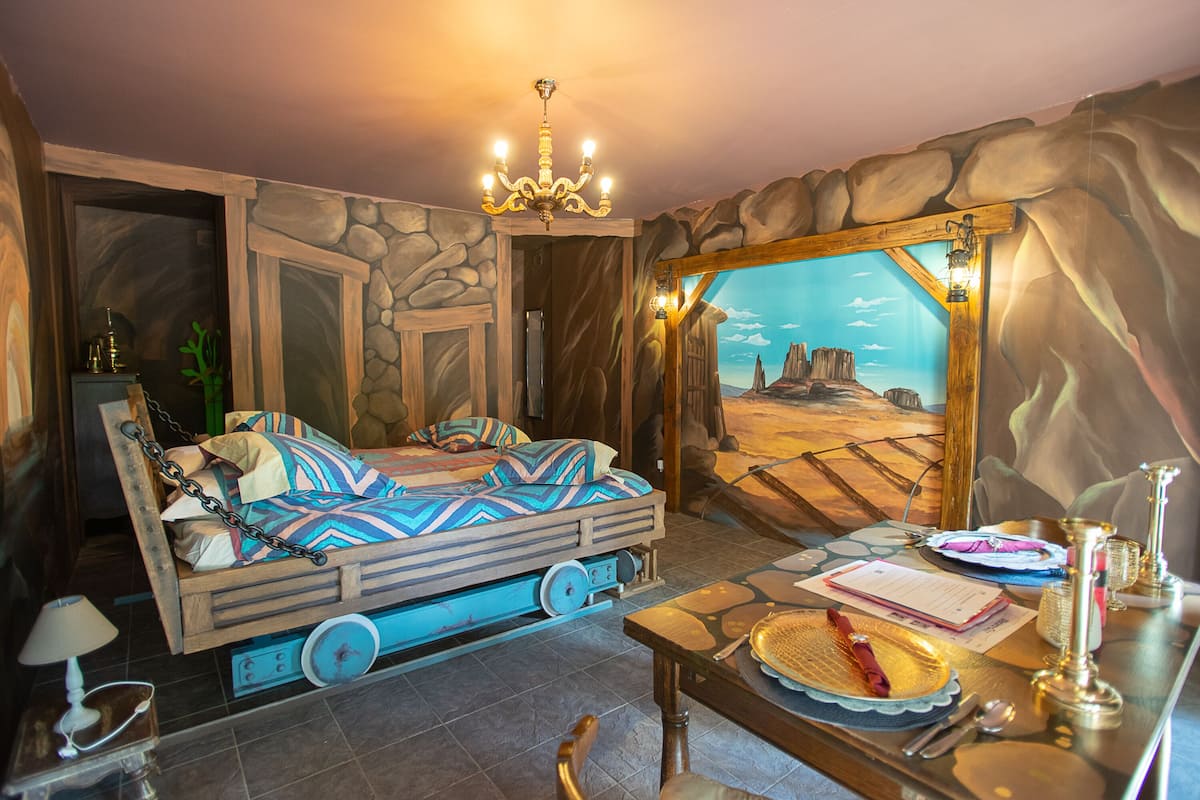 A room with a bed, a table with a plate and utensils, a chandelier, and a mural of a desert scene.