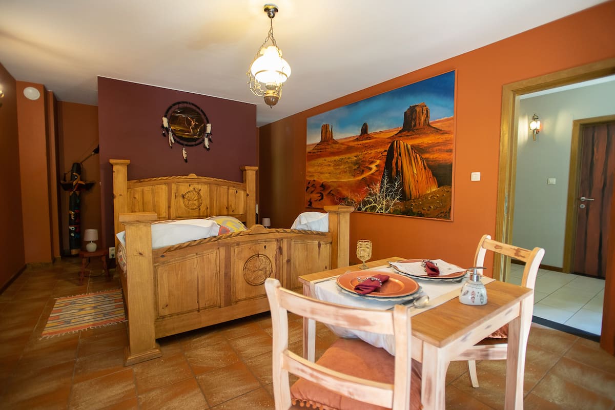 A room with a wooden bed, a dining table with chairs, and a painting of a desert landscape on the wall.