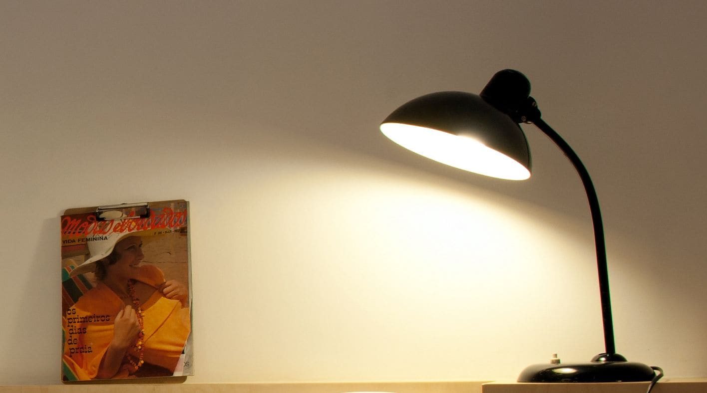 Eine Schreibtischlampe mit weißem Lampenschirm erhellt eine Wand.