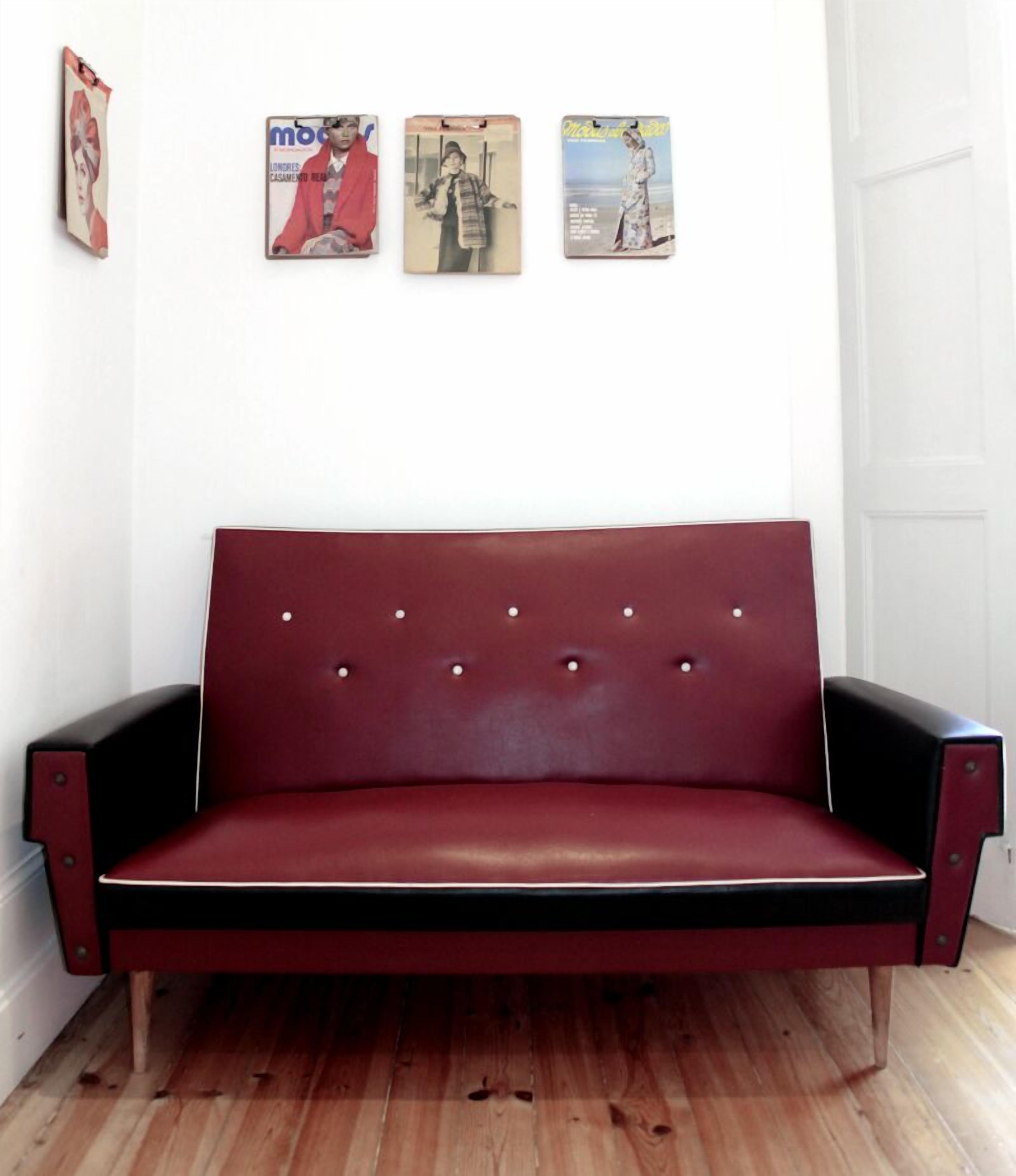 Un sofá de cuero rojo con un respaldo alto y detalles capitonados, colocado frente a una pared blanca con tres carteles enmarcados encima.