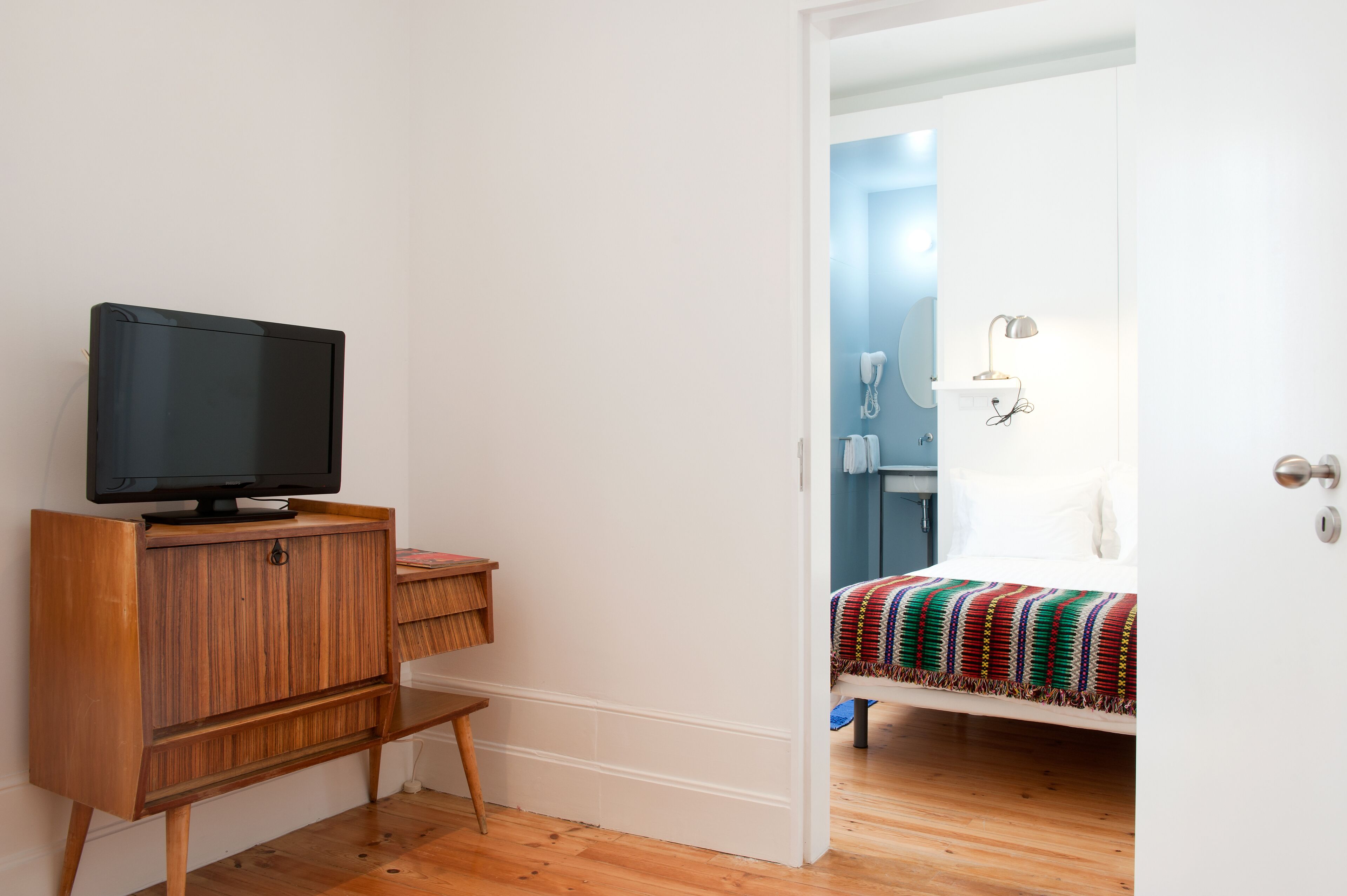 A room with a wooden TV stand, a bed with a colorful blanket, and a door leading to a bathroom with a mirror and a lamp.