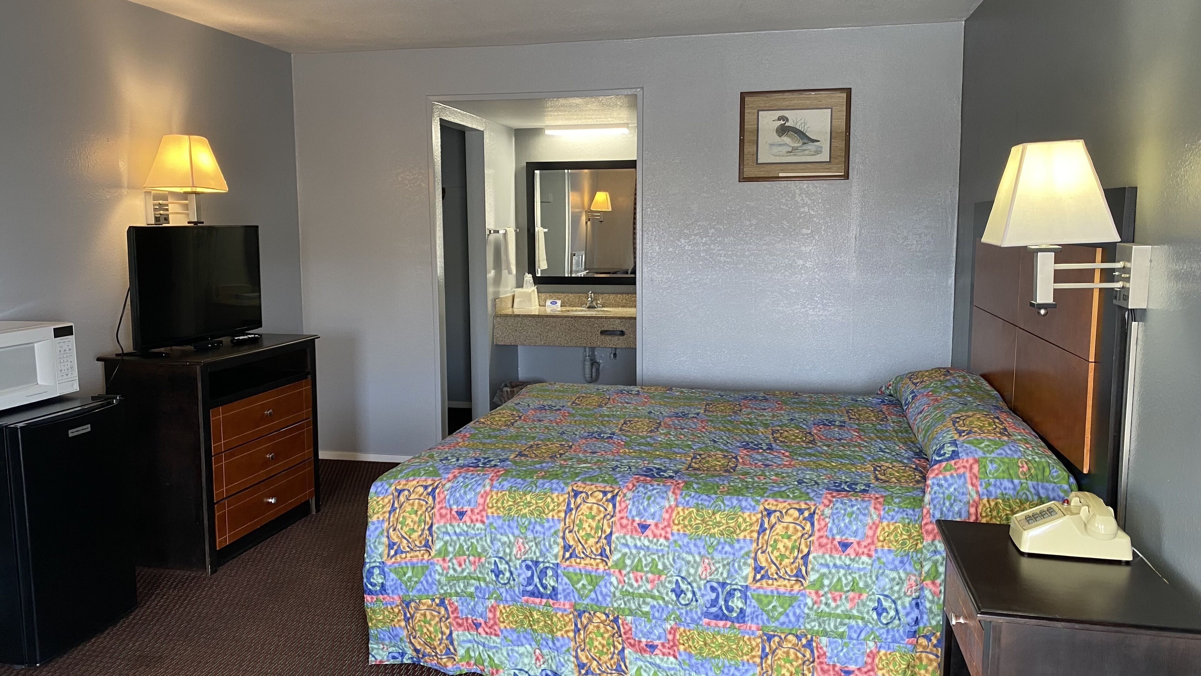 A hotel room with a bed, a dresser, a television, a lamp, and a bathroom visible through an open door.