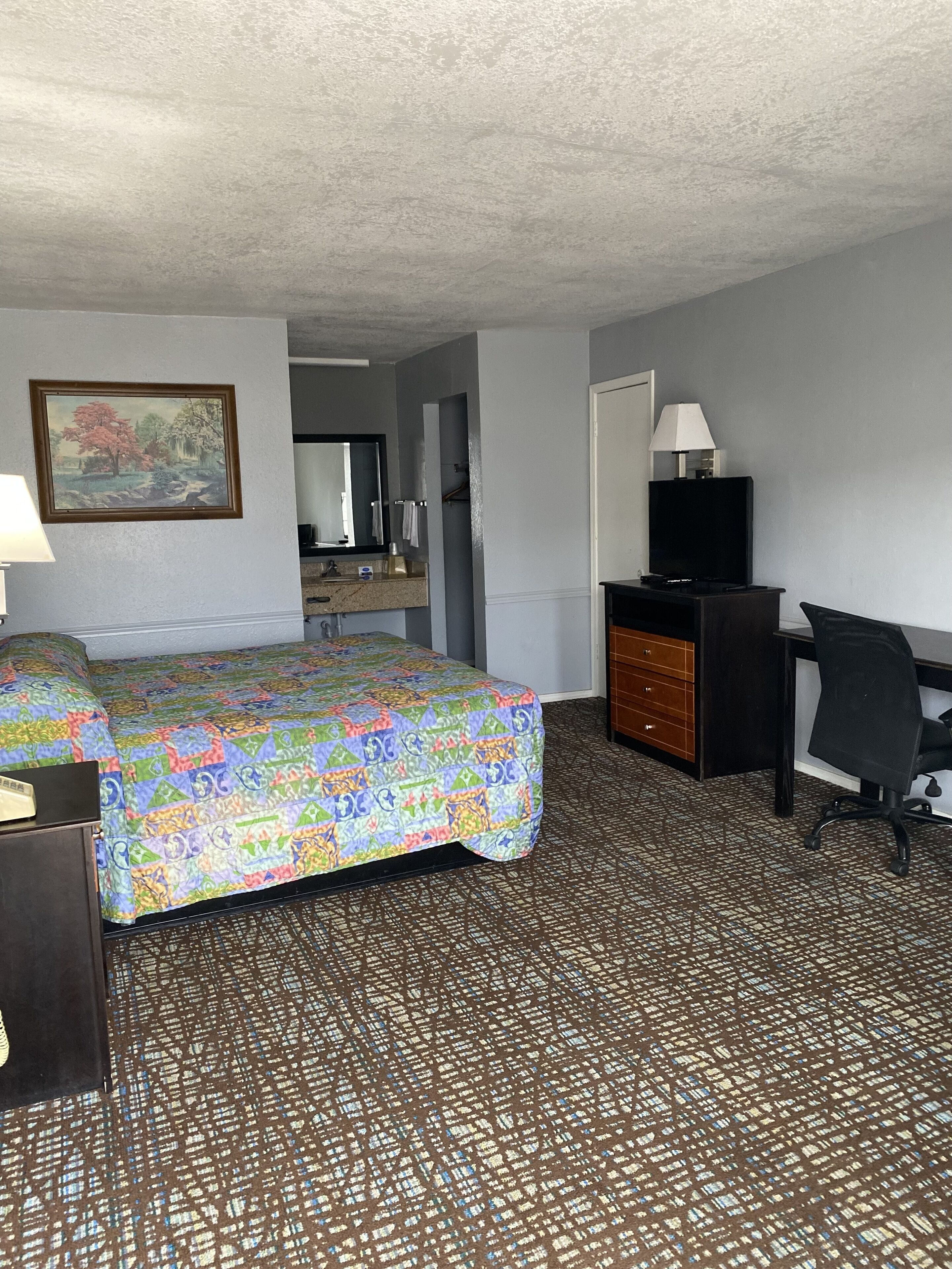 A hotel room with a bed, a desk, a chair, a dresser, a television, and a bathroom visible through an open door.