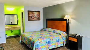 A hotel room with a bed, a nightstand with a lamp, a painting on the wall, and a bathroom visible through an open door. - Wilshire Inn (Cushing)