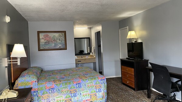 Una habitación de hotel con cama, escritorio y silla, televisor, cómoda y baño con lavamanos y espejo, además de un cuadro en la pared.