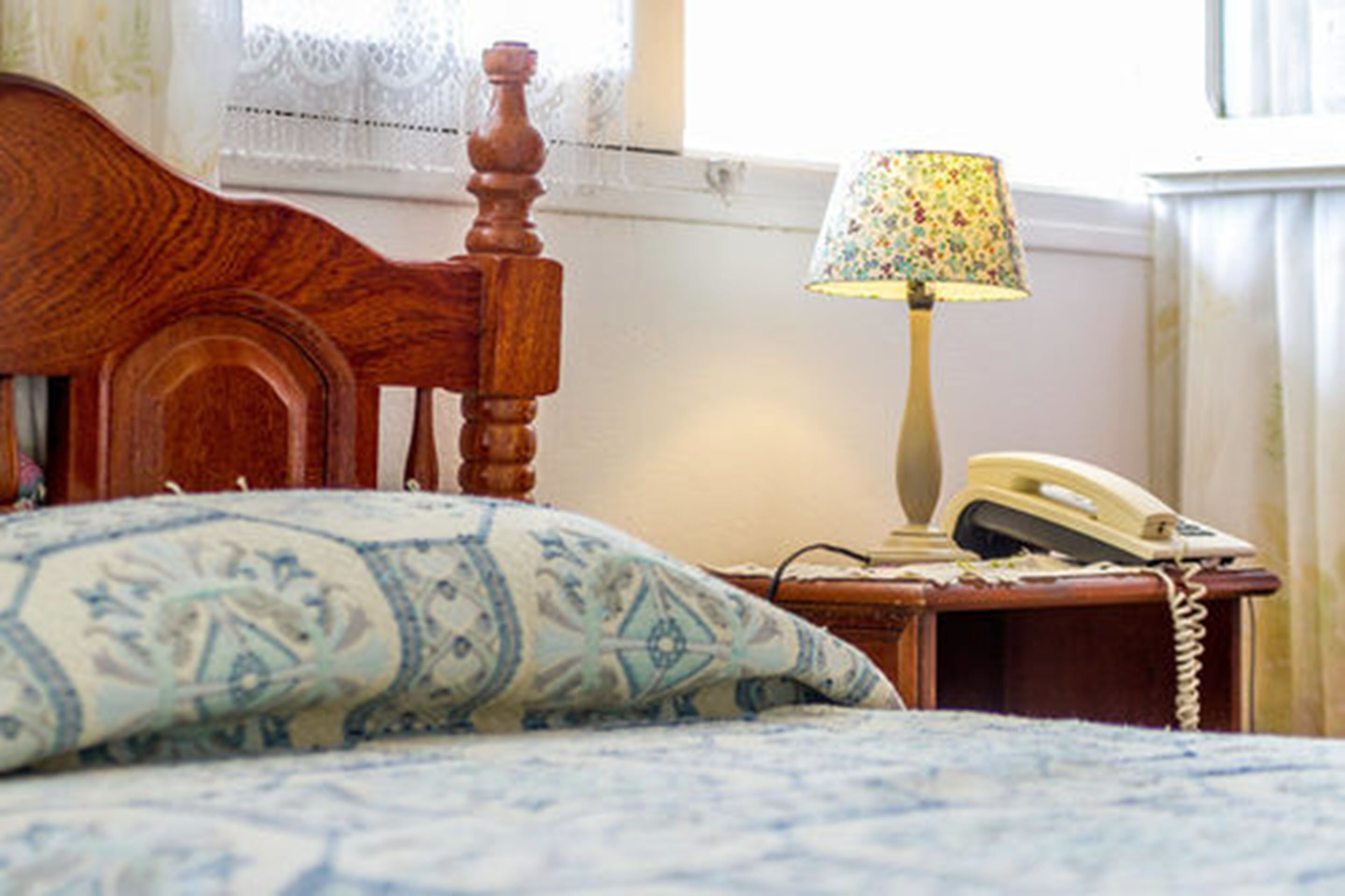 Una cama con un cojín estampado, un cabecero de madera, una lámpara con pantalla floral y un teléfono en la mesita de noche.