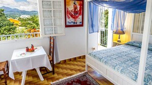 A bedroom with a bed, a table, a window with a view, and a painting on the wall. - Les chambres d'hotes de la Villa Cayol (Fort-de-France)