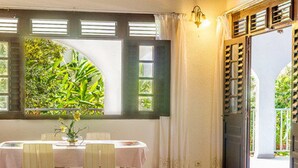 A dining area with a table set for two, a window with shutters, and a view of greenery outside. - Les chambres d'hotes de la Villa Cayol (Fort-de-France)