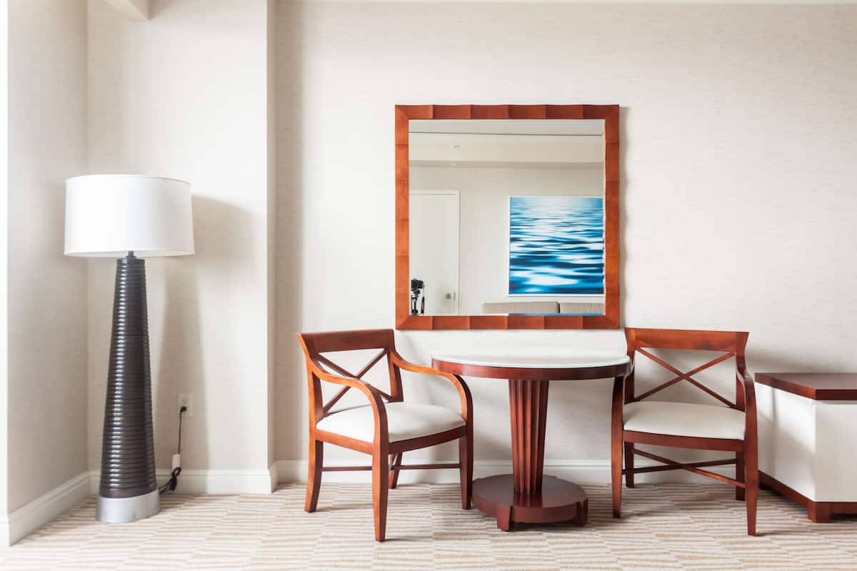 A room with a round table, two chairs, a lamp, and a large mirror with a painting.