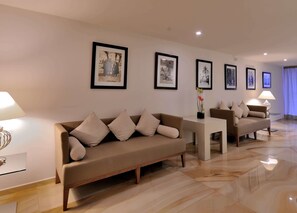 A modern living room with a beige sofa, a white coffee table, and a TV on a stand. There are framed pictures on the wall and a vase with flowers on the coffee table.