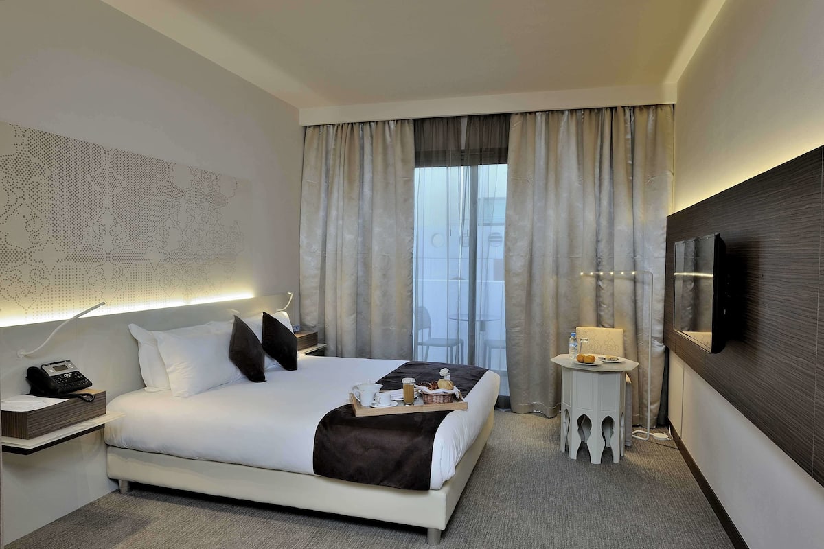 Une chambre d’hôtel avec un grand lit, une table de chevet avec une lampe, un téléviseur fixé au mur et une fenêtre avec des rideaux.