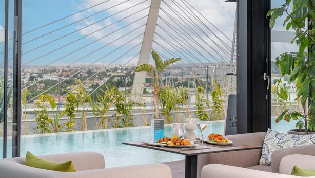 Een moderne hotelkamer met een groot raam dat uitkijkt op een stadsgezicht en een brug. De kamer heeft een zwembad, een eettafel met menukaart en comfortabele zitplaatsen.