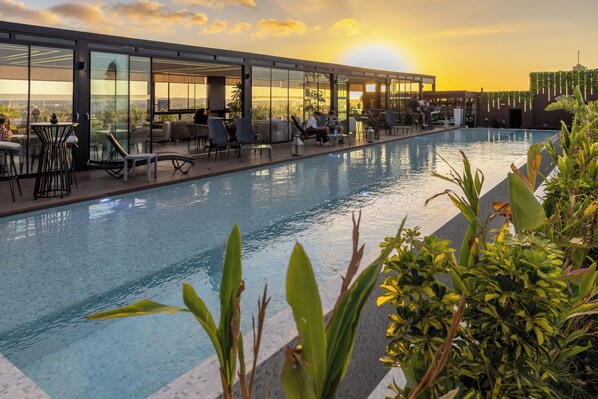 A rooftop pool with lounge chairs and tables, surrounded by glass walls and plants.