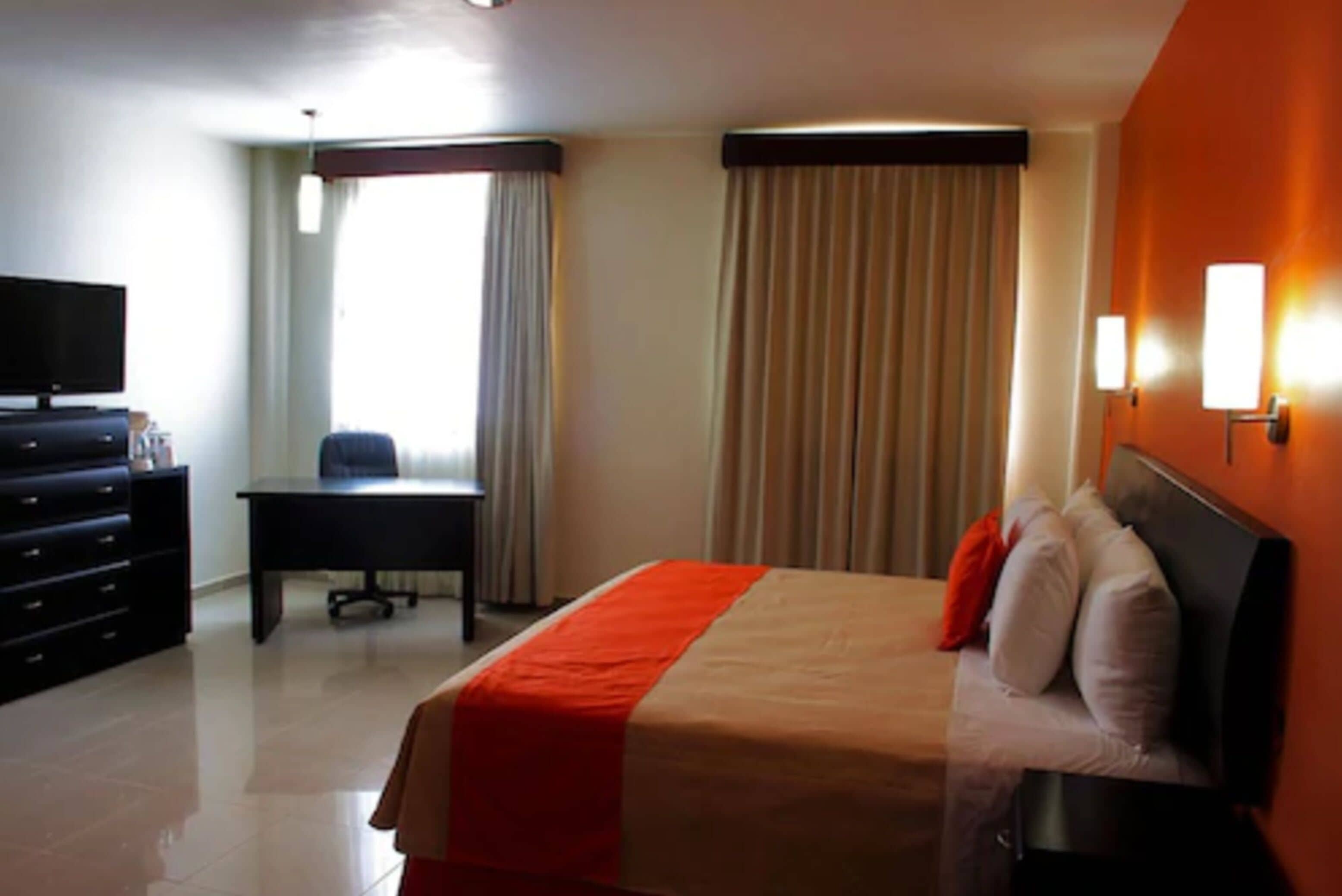 Una habitación de hotel con cama, escritorio, televisor y ventana con cortinas.