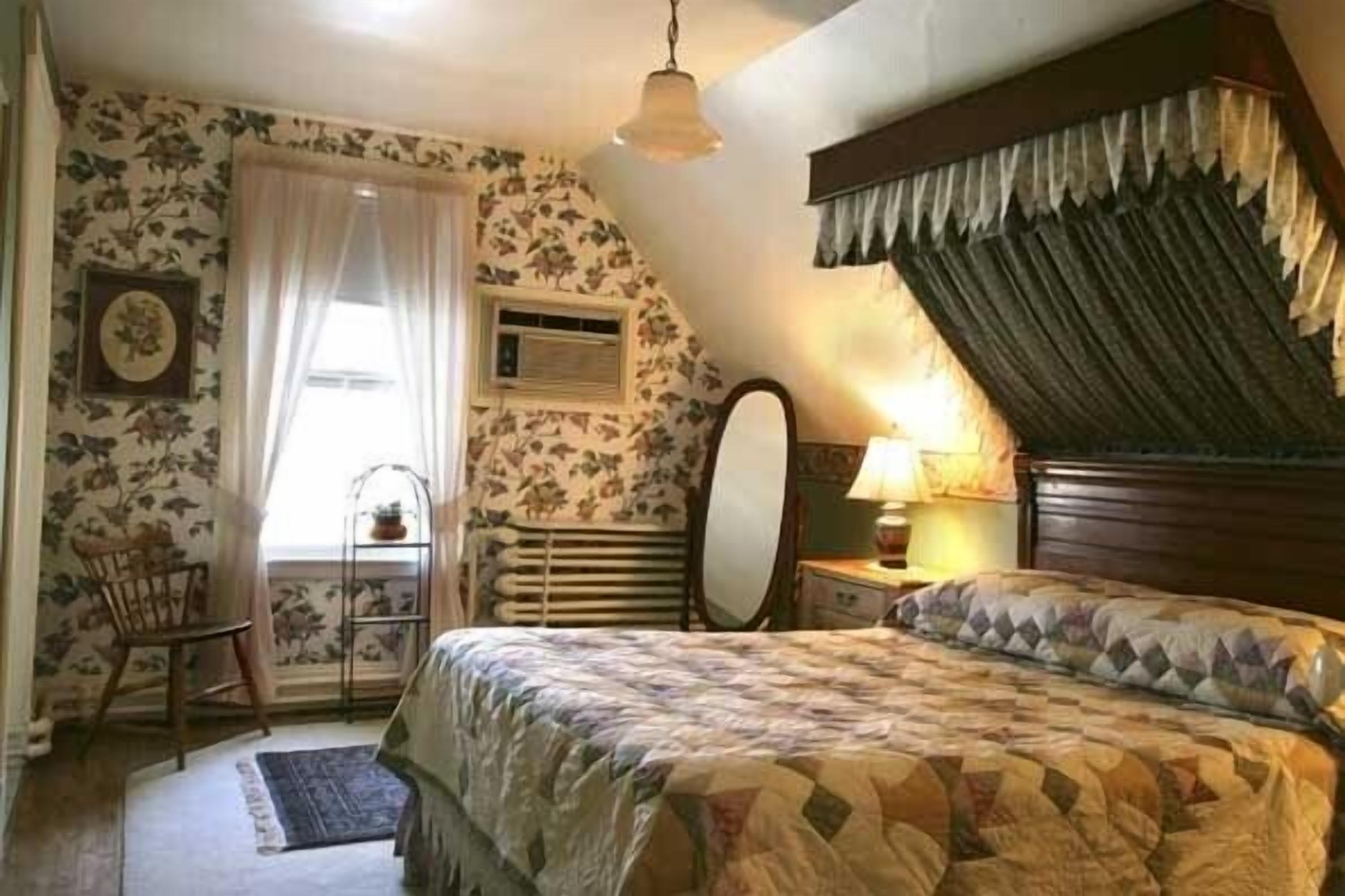 A bedroom with a bed, a chair, a window with curtains, a radiator, and a lamp.