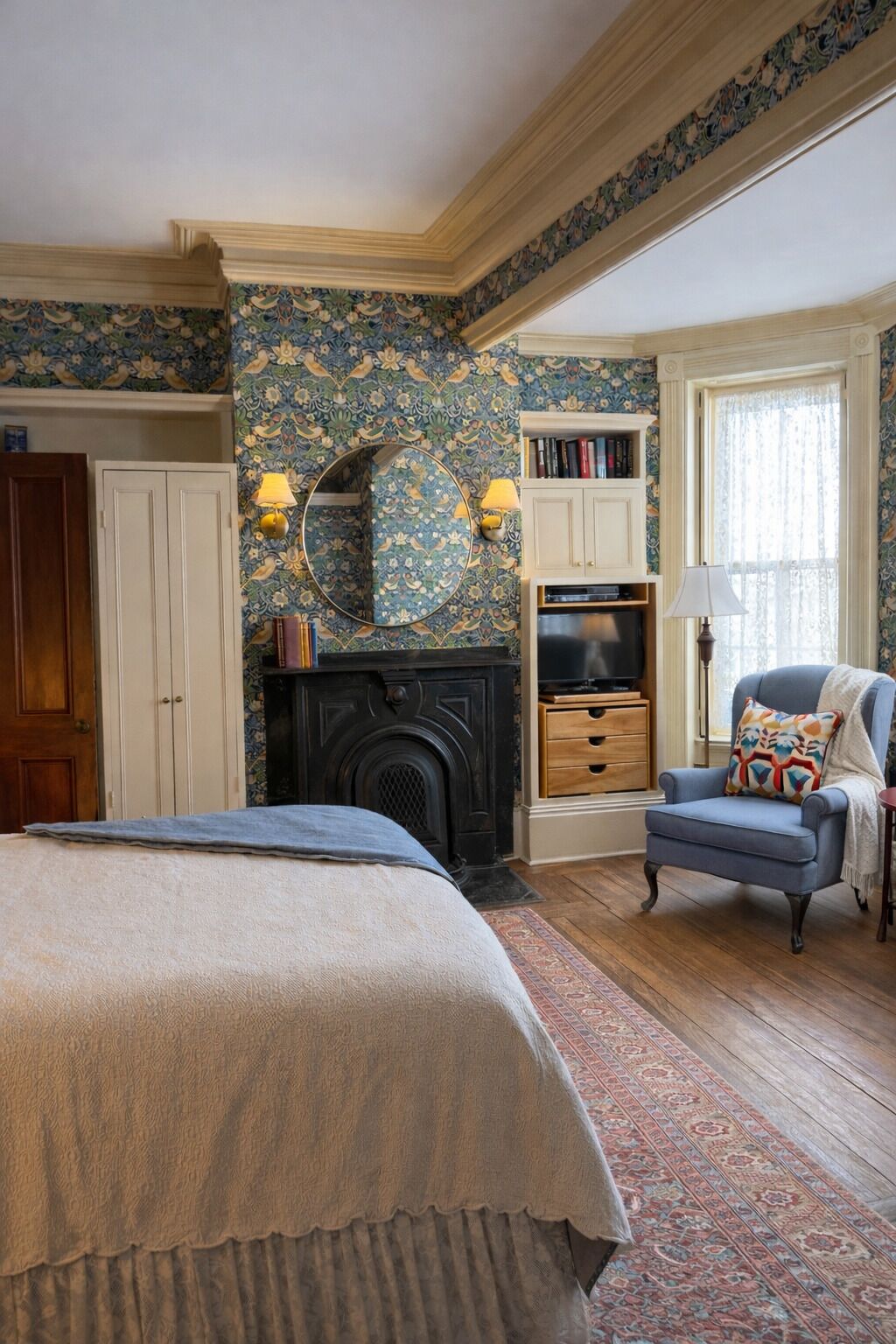 A bedroom with a bed, a fireplace, a blue armchair, and a bookshelf.