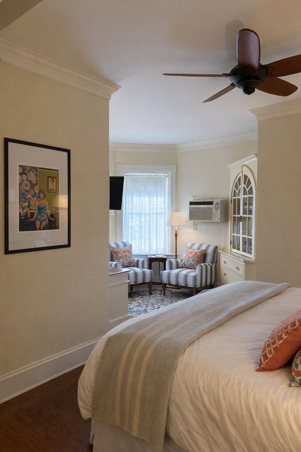 A bedroom with a bed, a ceiling fan, a window with curtains, a chair, a lamp, and a framed picture on the wall.
