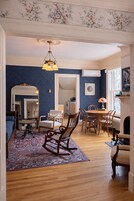 Habitación con pared azul, suelo de madera, una mesa de comedor con sillas, una mecedora, alfombra estampada, un espejo y una lámpara colgante.